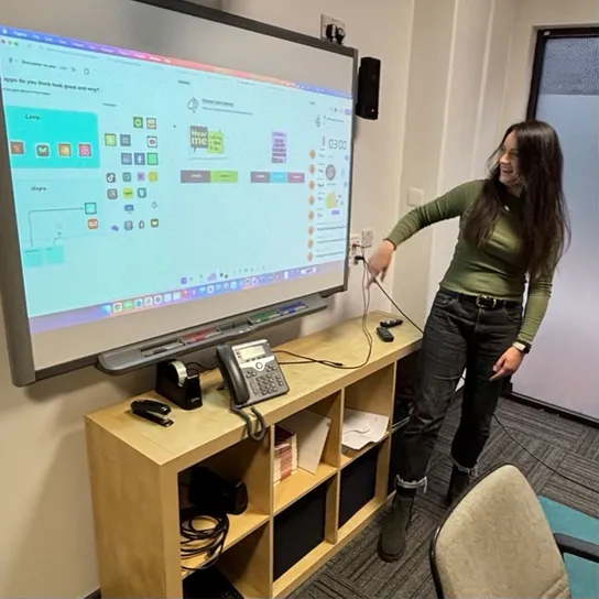 Smiling woman points and looks at large screen behind her, her body faces the room. The screen displays app icons and colour palettes.